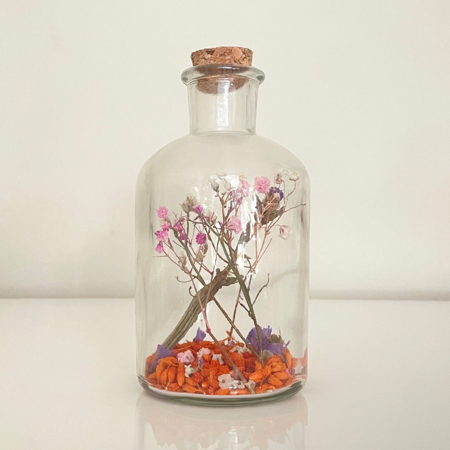 Glass bottle with a cork lid containing a pink, purple and orange dried flowers on a white surface.