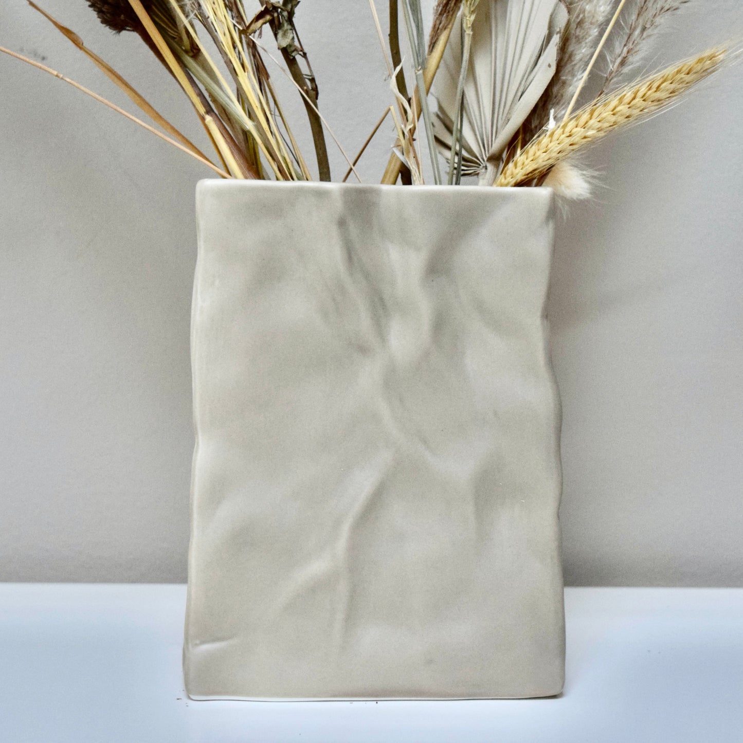 Beige ceramic vase with dried flowers n a white surface.