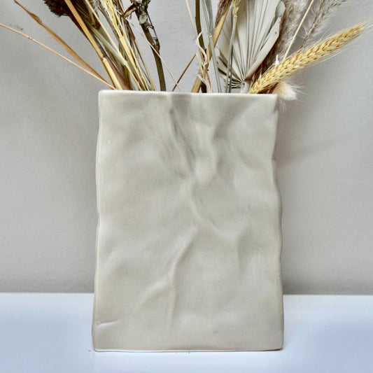 Beige ceramic vase with dried flowers n a white surface.