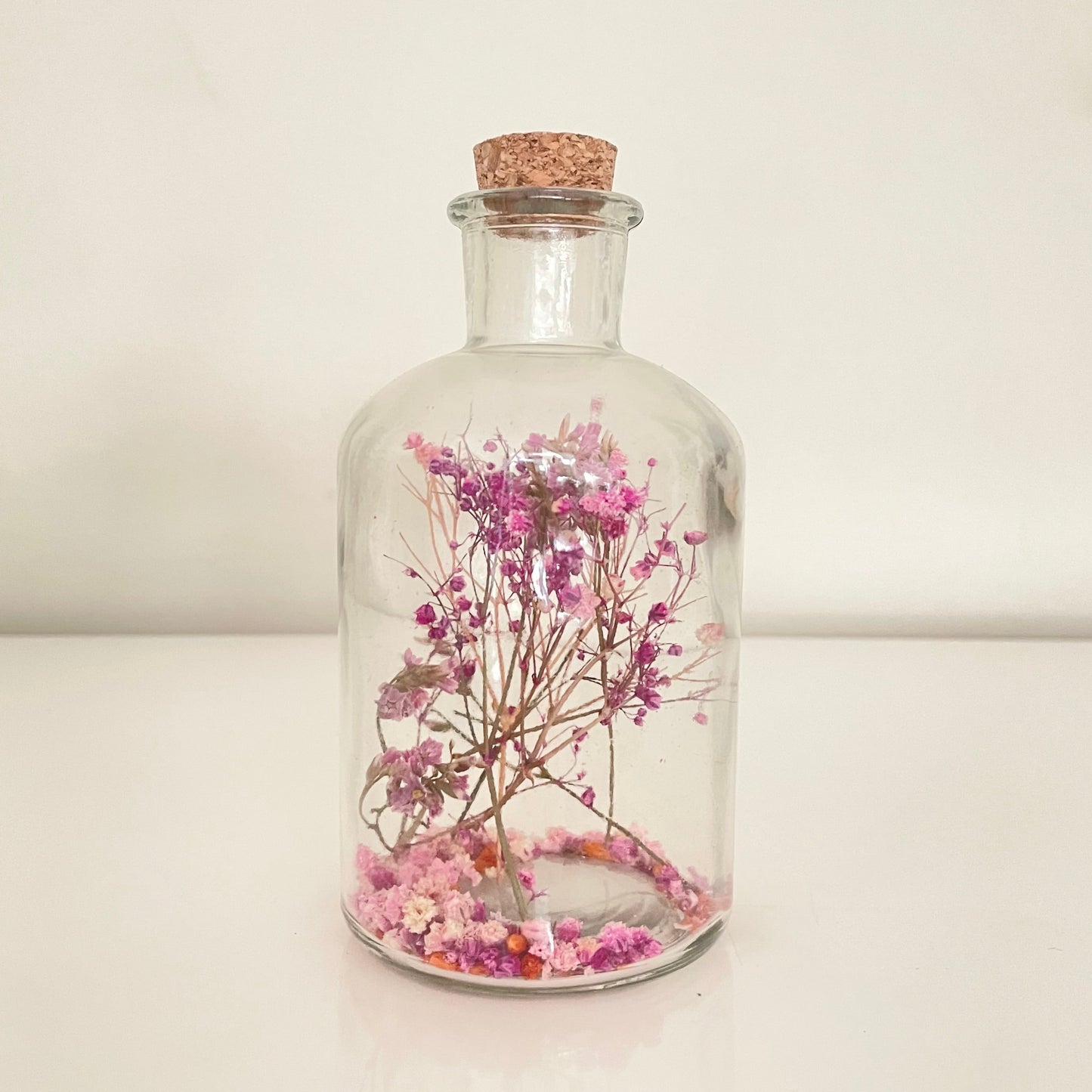 Glass bottle with cork lid containing pink and purple dried flowers on a light background.