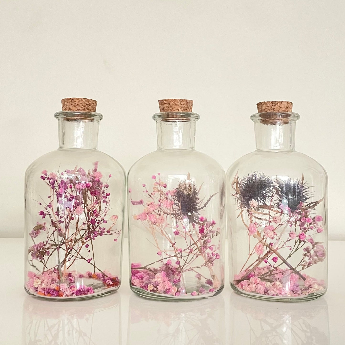 Glass bottles with cork lid containing pink, purple and blue dried flowers on a light background.