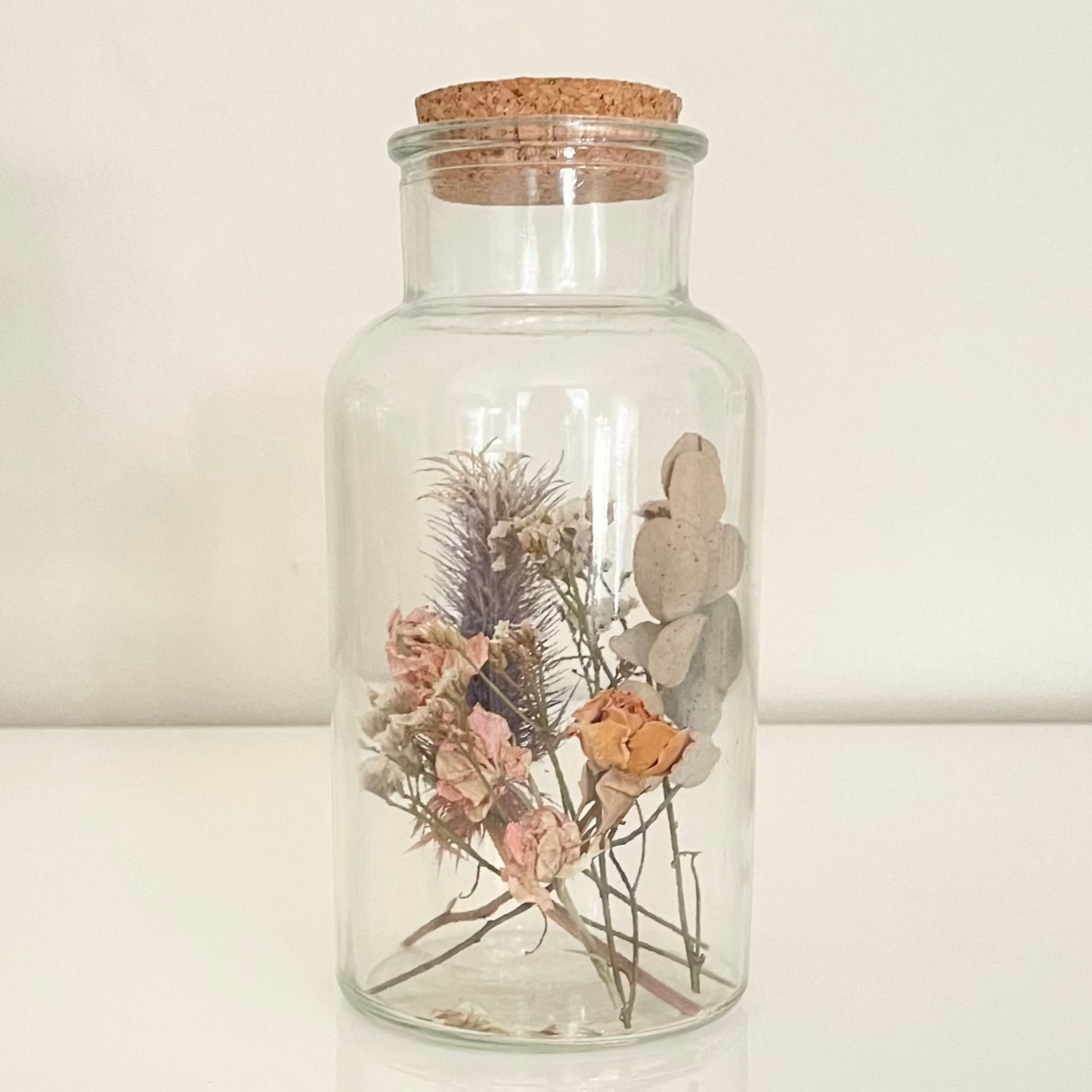 Glass bottle filled with yellow, pink and green dried flowers on a white background.