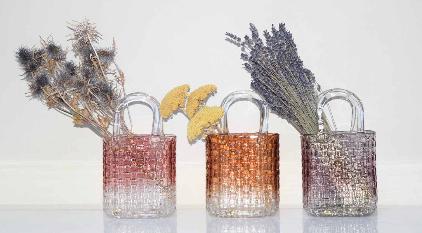 Three glass vases in pink, amber and purple with woven designs, each containing different dried plants on a white background.