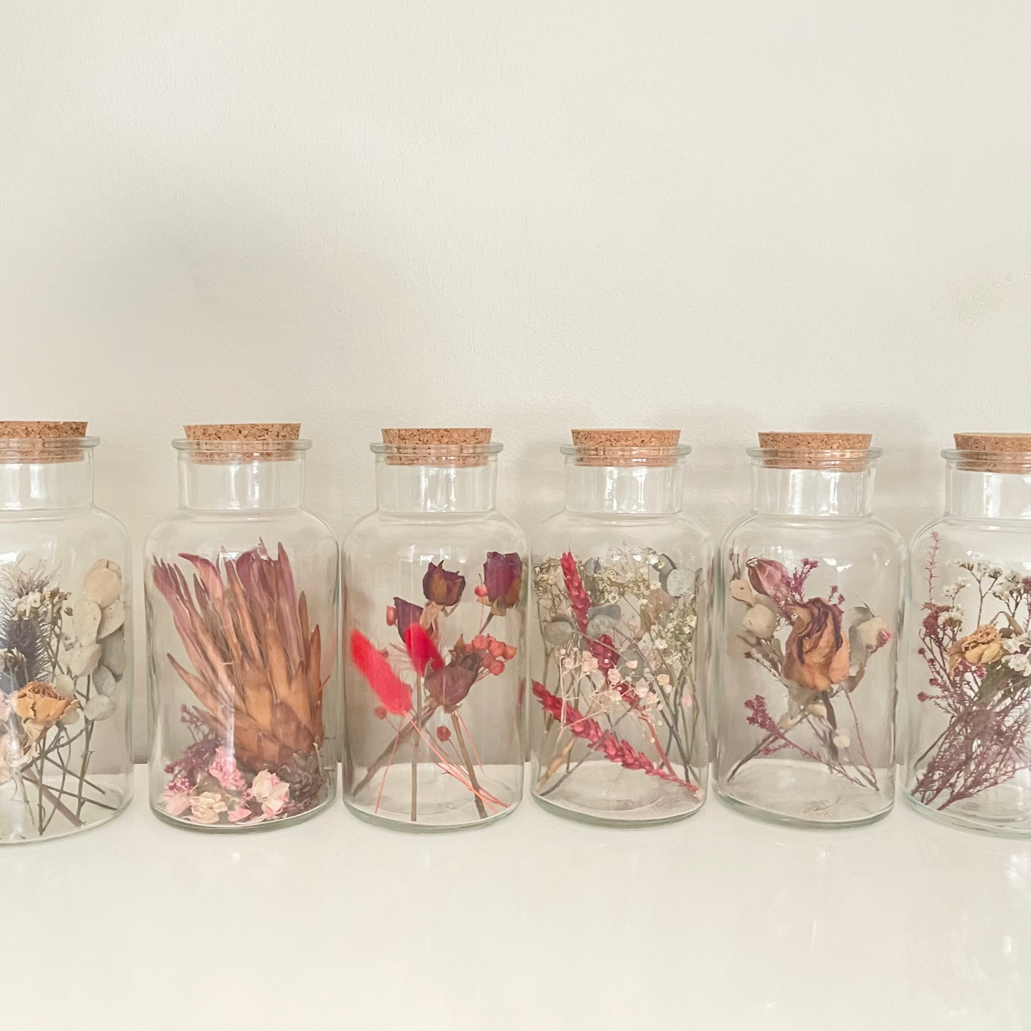 Glass bottles with cork lids containing dried floral arrangements on a white background.
