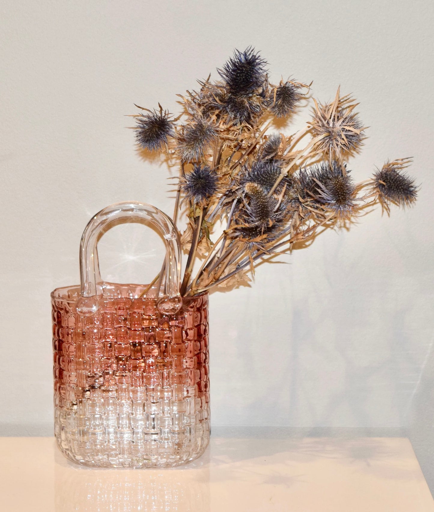 Decorative pink glass vase with a woven design holding dried flowers on a white background.