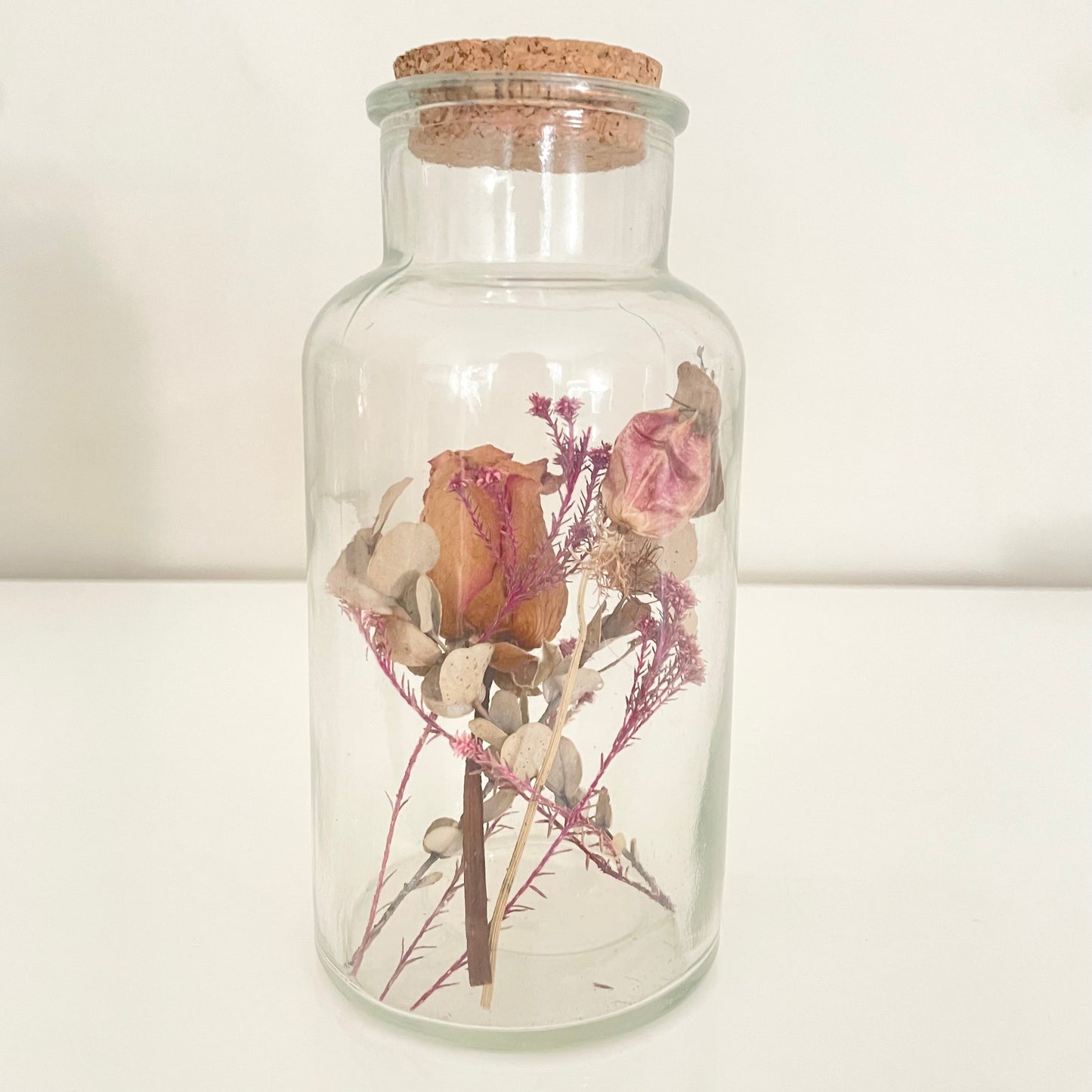 Glass bottle with cork lid containing dried flowers on a light background.