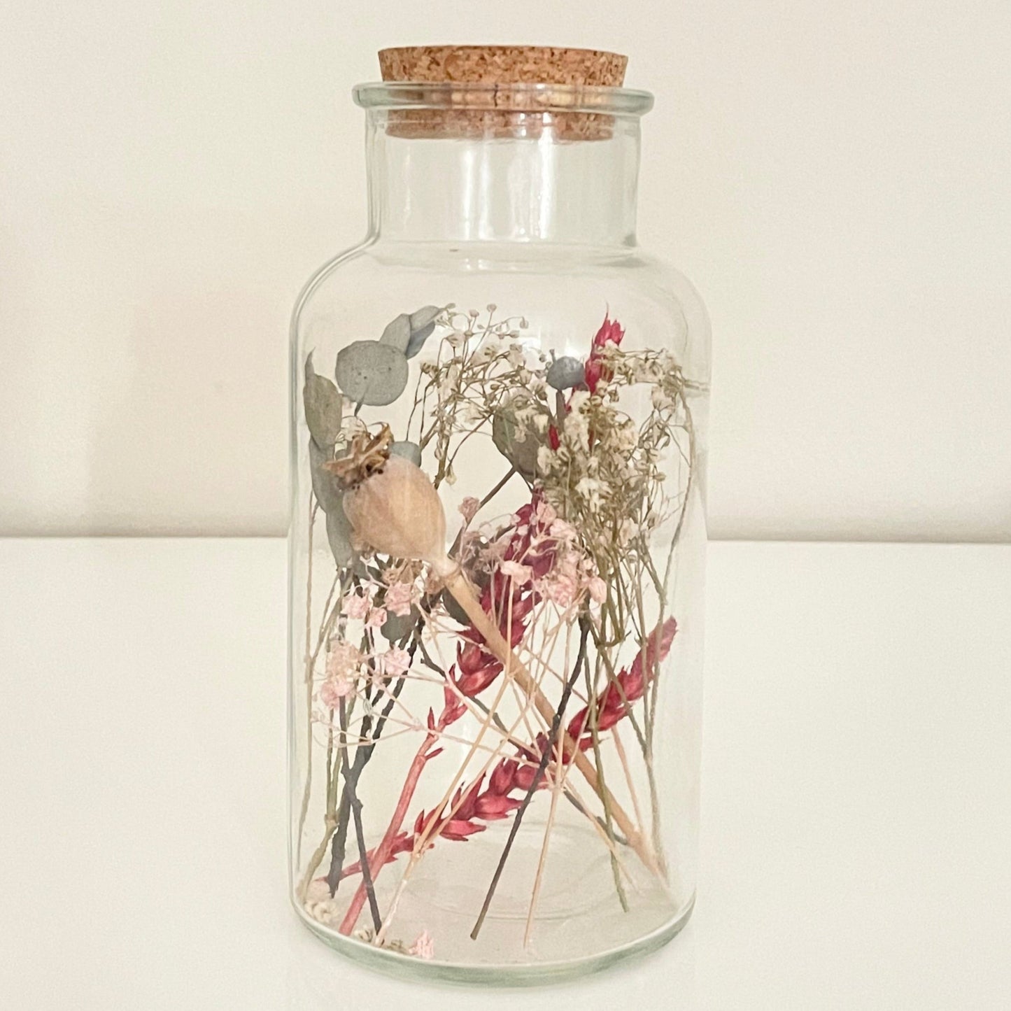 Glass bottle with cork lid containing dried flowers on a light background.