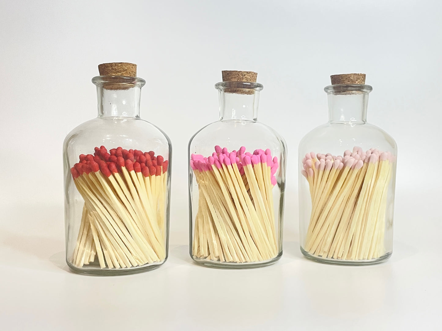 Three glass bottles with cork lids containing matches with pink tops on a white background. 