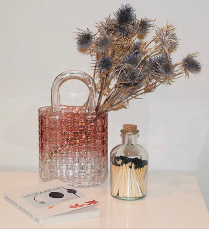 Decorative scene with a woven pink glass  bag vase, dried flowers, and our bottled apothecary safety matches on a white surface.