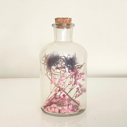 Glass bottle with cork lid containing pink, purple and blue dried flowers on a light background.