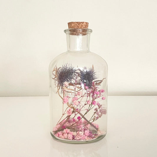 Glass bottle with cork lid containing pink, purple and blue dried flowers on a light background.