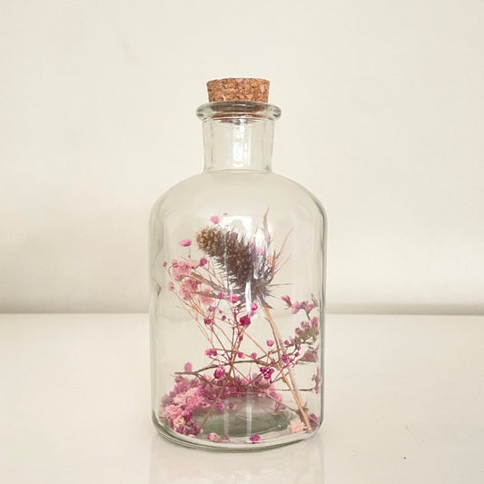 Glass bottle with cork lid containing pink, purple and blue dried flowers on a light background.