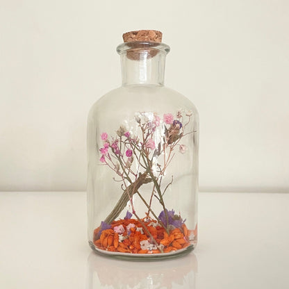 Glass bottle with a cork lid containing a pink, purple and orange dried flowers on a white surface.