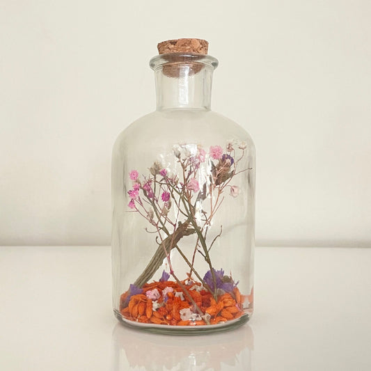 Glass bottle with a cork lid containing a pink, purple and orange dried flowers on a white surface.