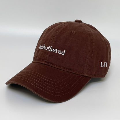 100% Brown washed cotton cap with 'unbothered' white embroidery with side emblem 'un'. 