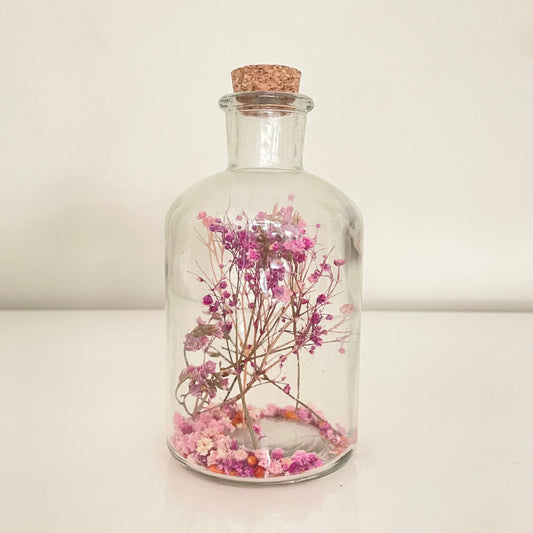 Glass bottle with cork lid containing pink and purple dried flowers on a light background.