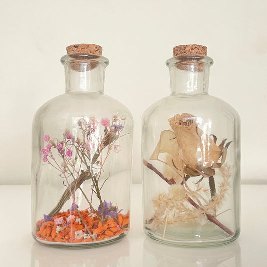 Glass bottles with cork lids containing dried flowers on a white surface.