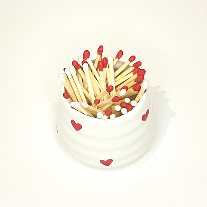 White ceramic holder/cup with heart designs filled with red and white matches on a white background.