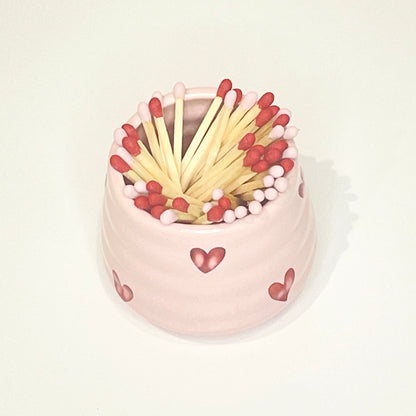 Pink ceramic holder/cup with heart designs filled with red and light pink matches on a white background.