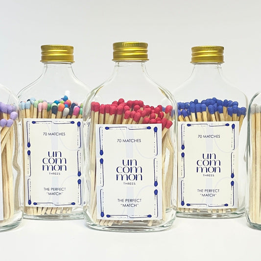Five glass bottles with gold lids filled with colourful long safety matches on a white background.