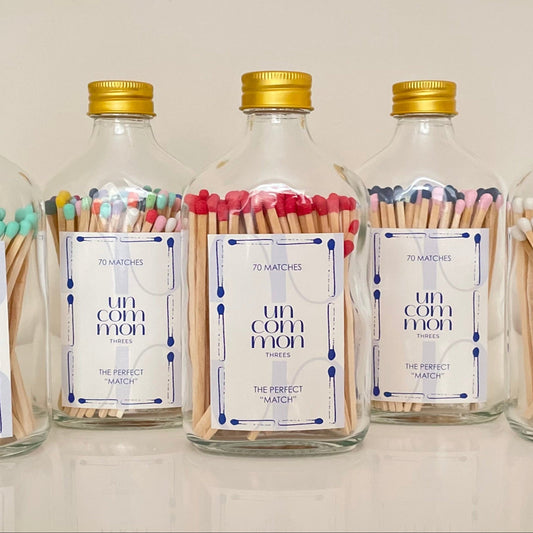 Five glass bottles with gold lids filled with colourful matches on a white background.