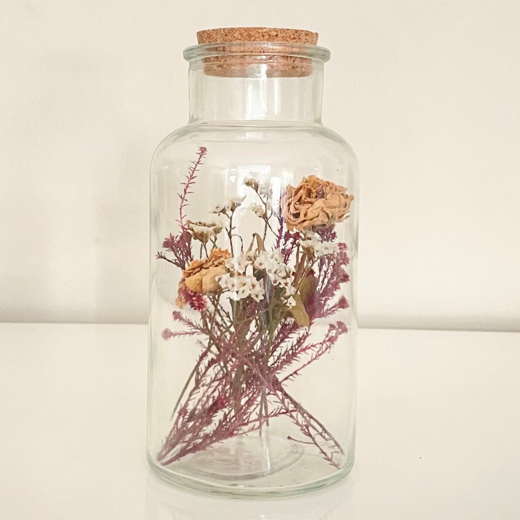 Glass bottle with cork lid containing dried floral arrangements on a white background.