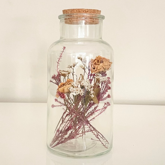 Glass bottle with cork lid containing dried floral arrangements on a white background.