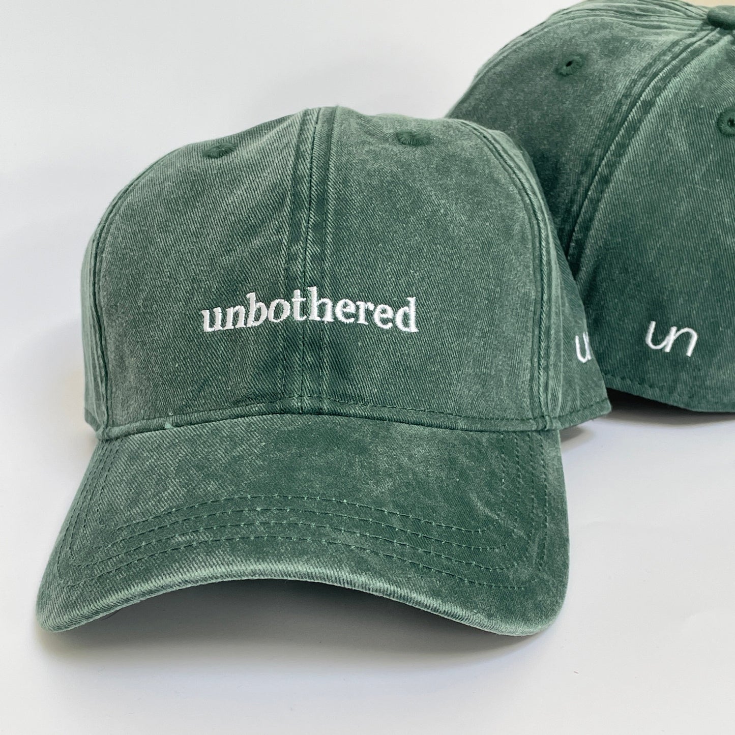 100% Green washed cotton cap with 'unbothered' white embroidery with side emblem 'un'. 