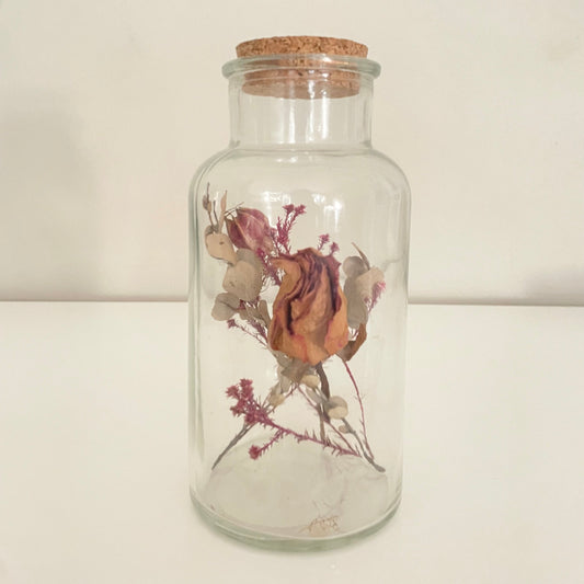 Glass bottle with cork lid containing dried flowers on a light background.