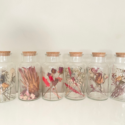 Glass bottles with cork lids containing dried floral arrangements on a white background.