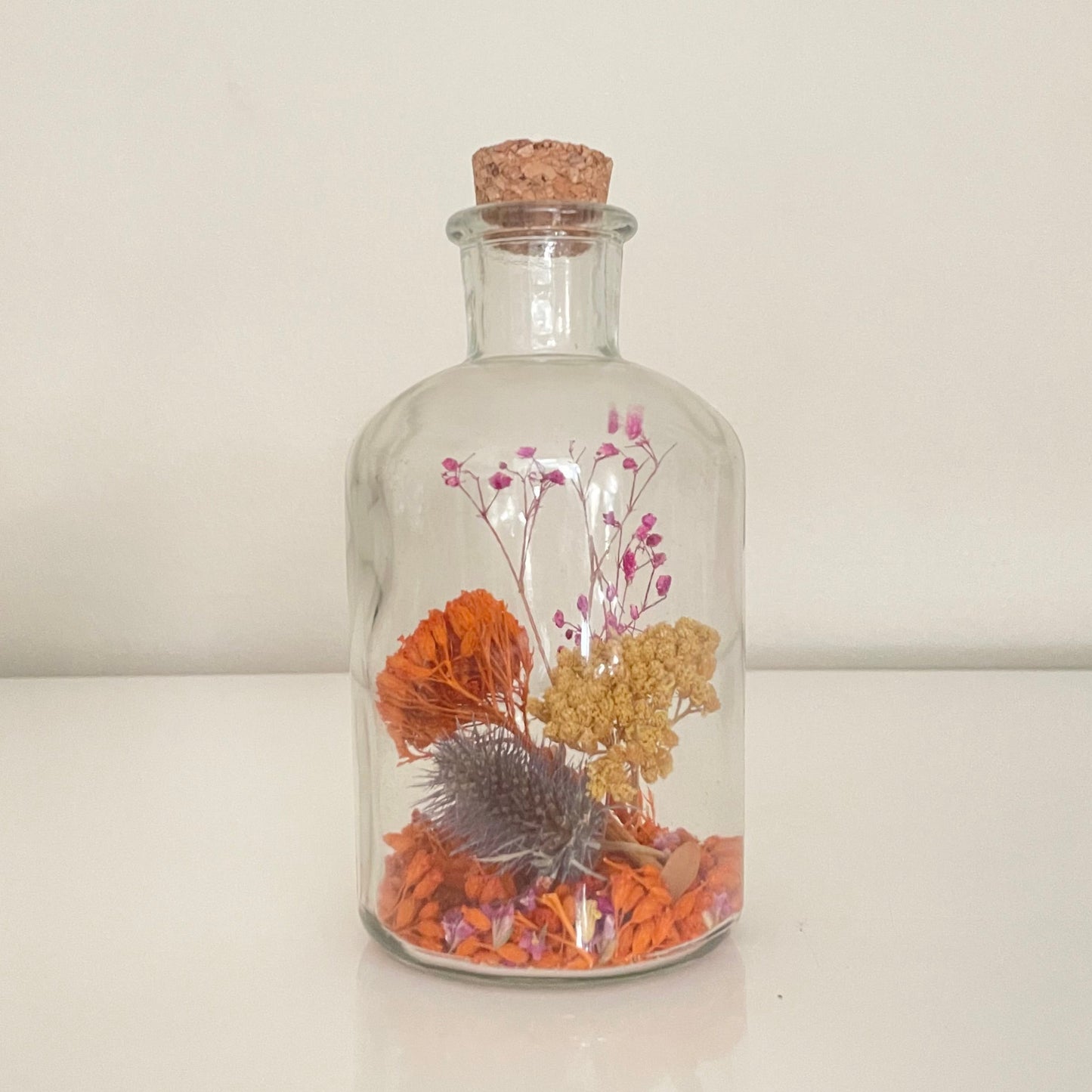 Glass bottle filled with yellow, orange and purple dried flowers on a white background. 