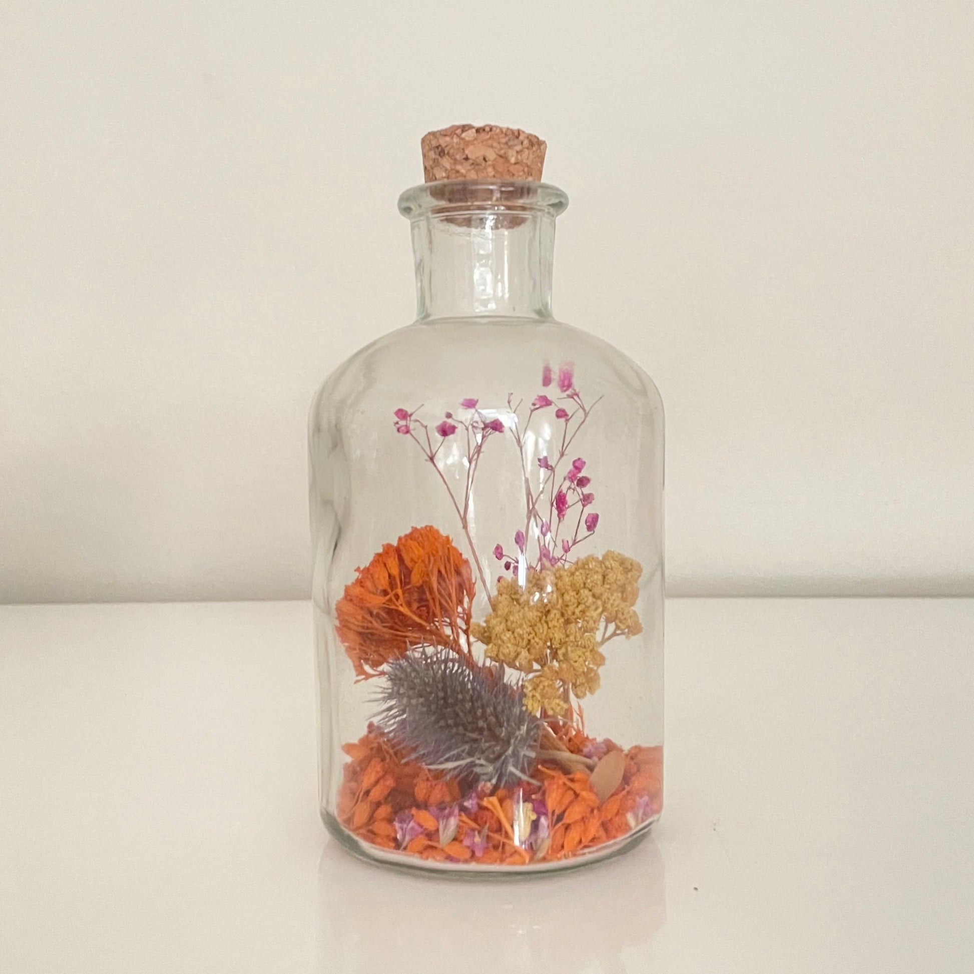 Glass bottle filled with yellow, orange and purple dried flowers on a white background. 