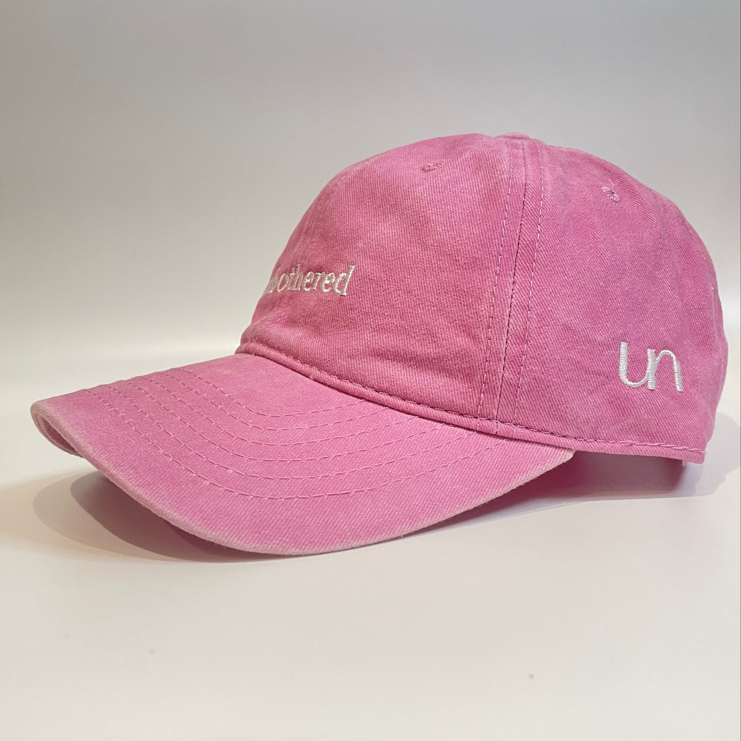 100% Pink washed cotton cap with 'unbothered' white embroidery with side emblem 'un'. 