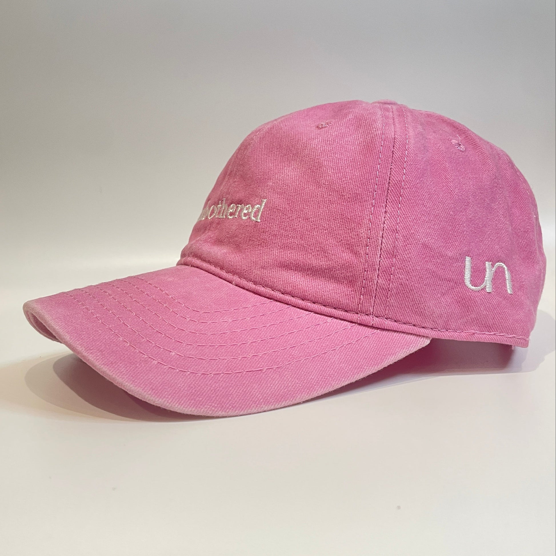 100% Pink washed cotton cap with 'unbothered' white embroidery with side emblem 'un'. 