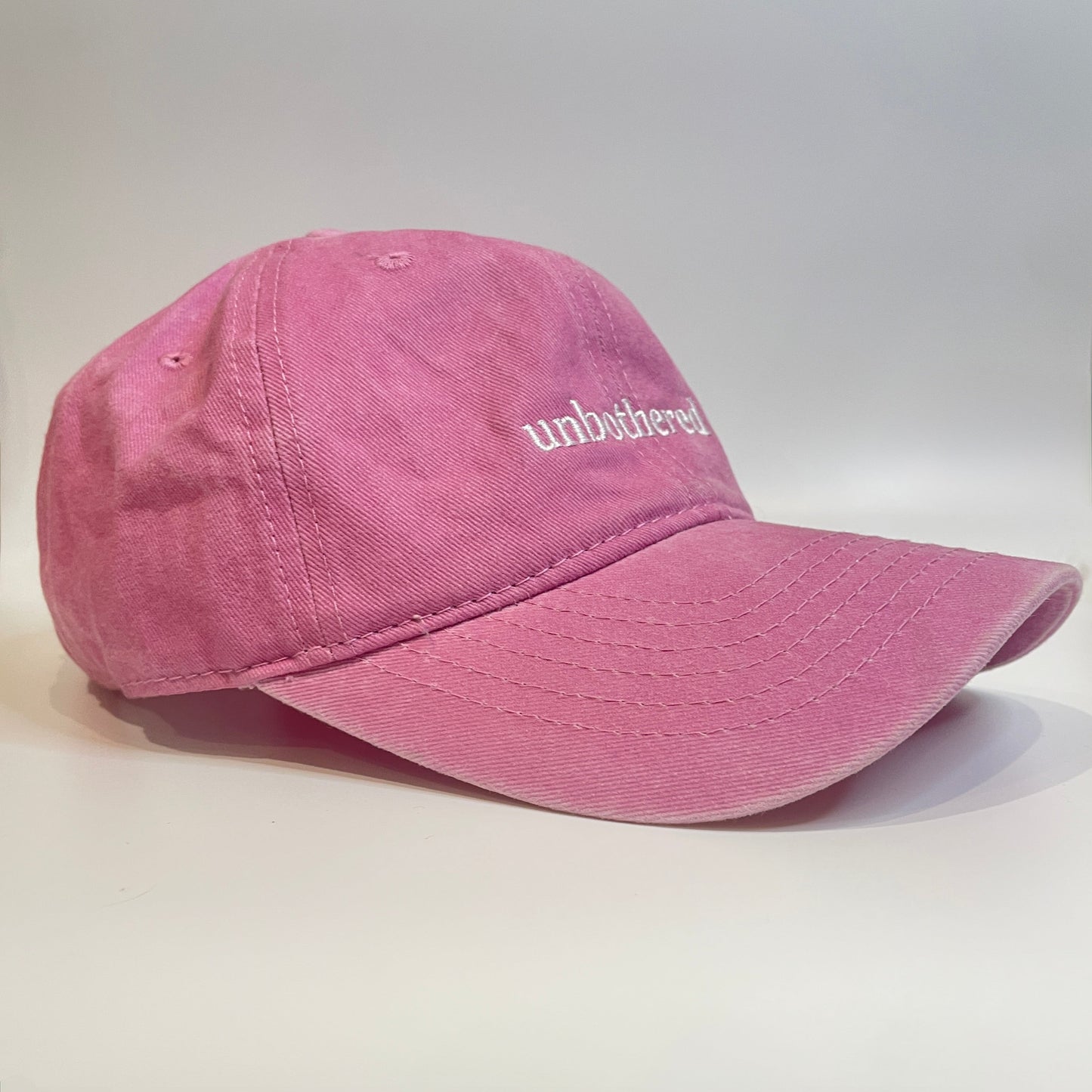 100% Pink washed cotton cap with 'unbothered' white embroidery with side emblem 'un'. 