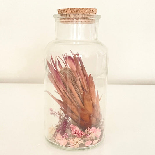 Glass bottle with cork lid containing dried flowers on a light background.