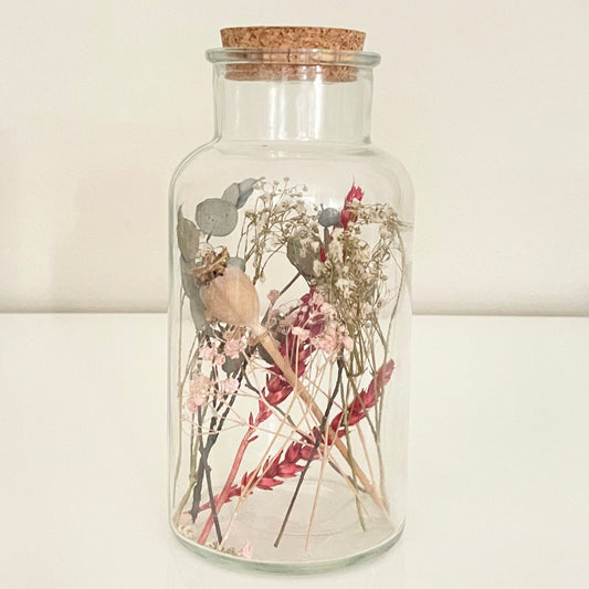 Glass bottle with cork lid containing dried flowers on a light background.