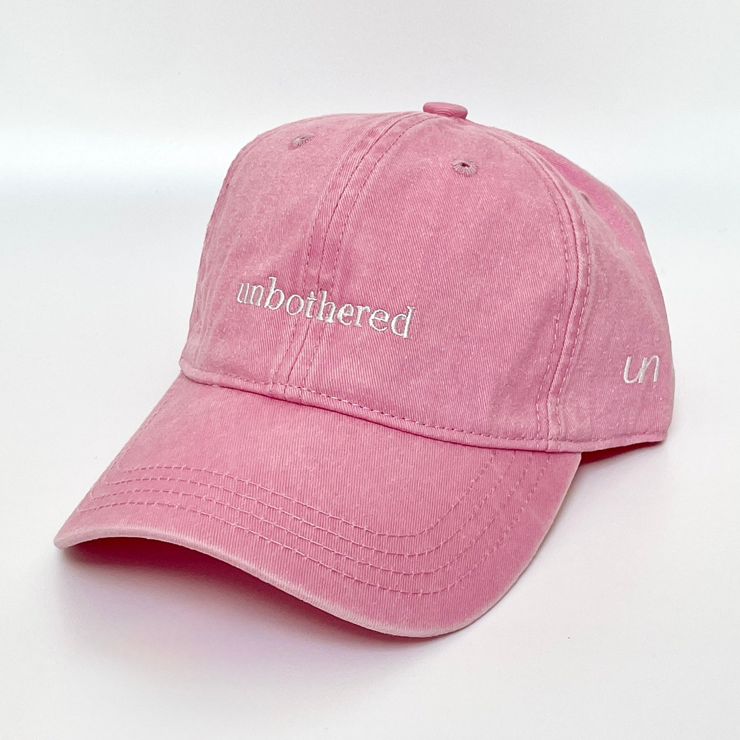 100% Pink washed cotton cap with 'unbothered' white embroidery with side emblem 'un'. 