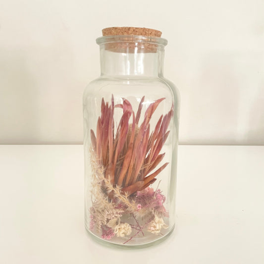 Glass bottle with cork lid containing dried flowers on a light background.