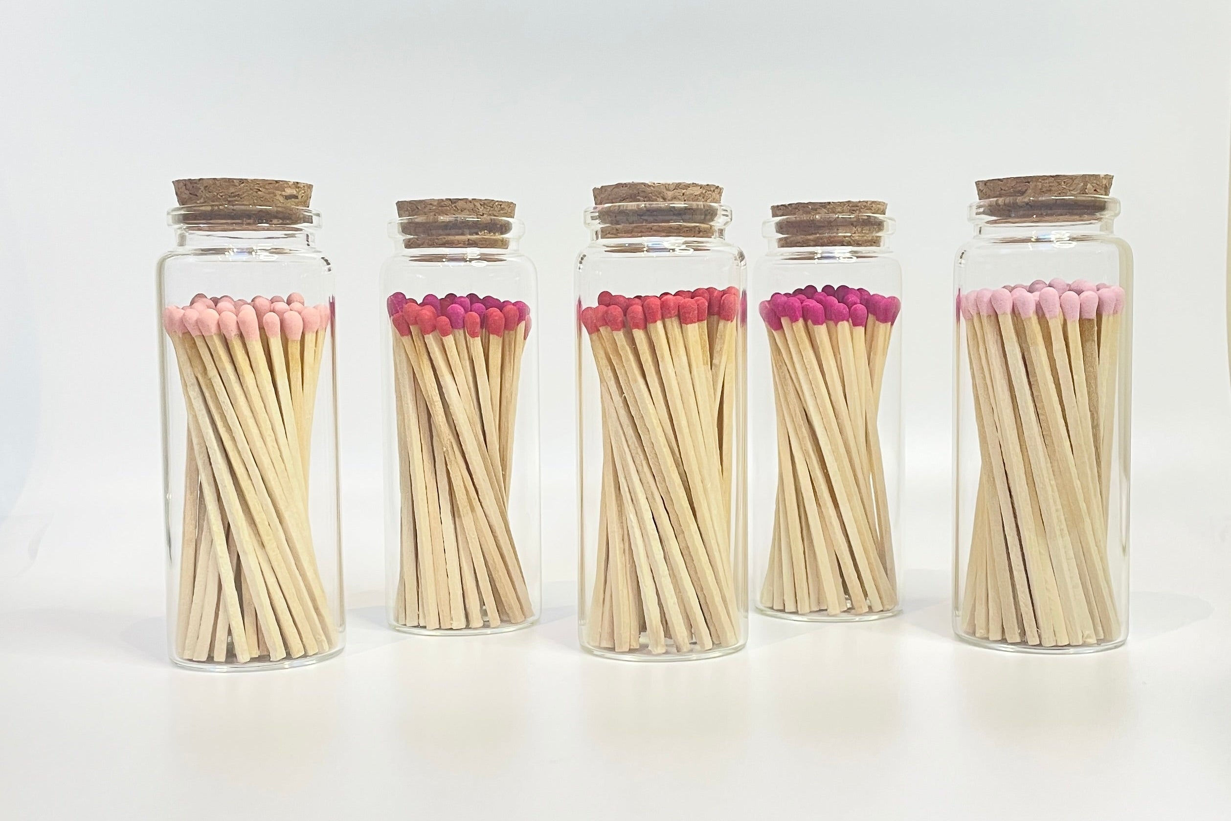 Five glass jars with cork lids filled with shades of pink matches on a white background