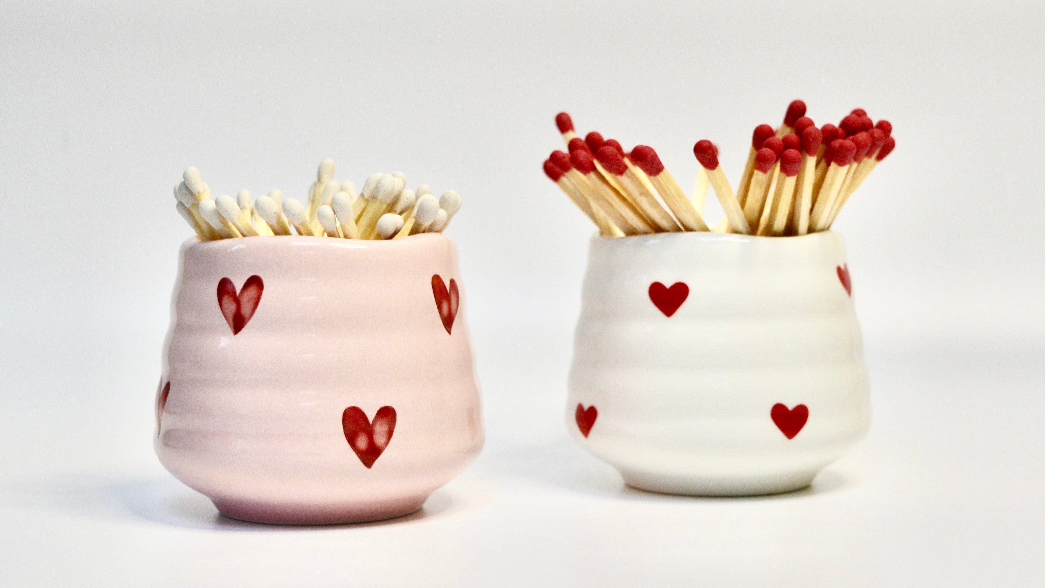 Two ceramic match holders with red hearts containing matches on a white background