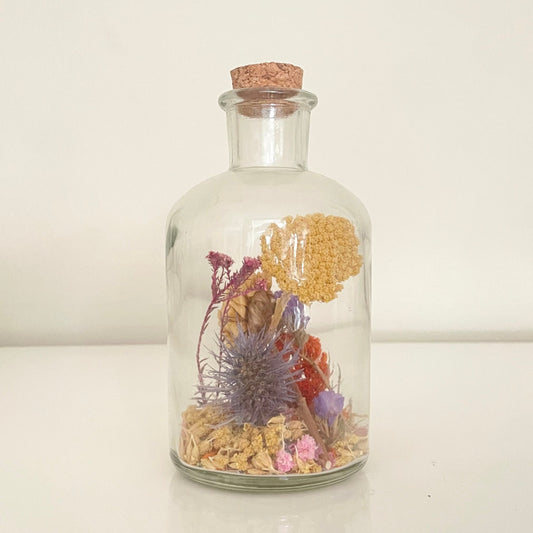 Glass bottle filled with yellow, orange and purple dried flowers on a white background. 